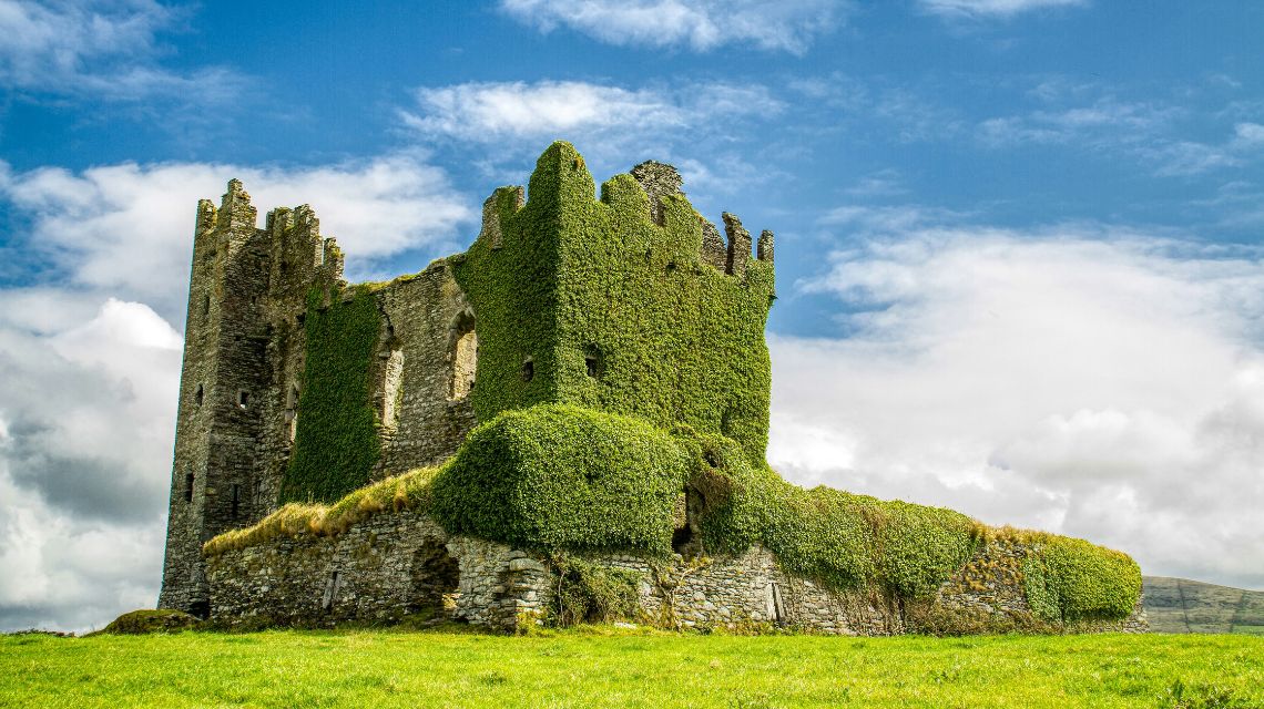 A historical stone castle ruin covered in lush green ivy stands against a bright blue sky with scattered clouds, evoking a sense of mystery and timelessness.