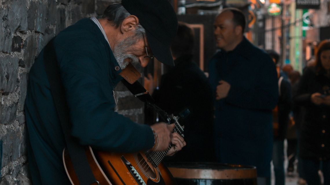 Elderly man in a hat strums a guitar against a stone wall on a lively street. Passersby walk by, some smiling, creating a warm, communal atmosphere.