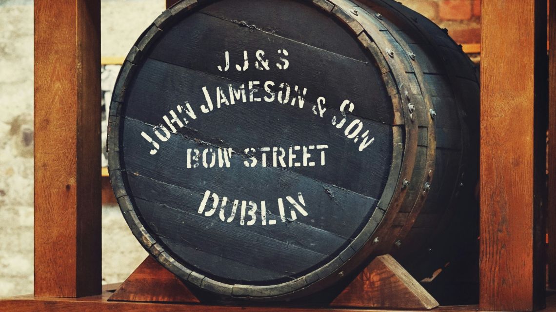 A wooden barrel with white text: "J.J.&S John Jameson & Son, Bow Street, Dublin." It rests between wooden supports, giving a rustic, historic feel.