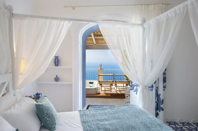 Bright, airy bedroom with sheer curtains, white and blue decor. Open archway reveals a sunny terrace with ocean view, lounge chairs, and serene atmosphere.