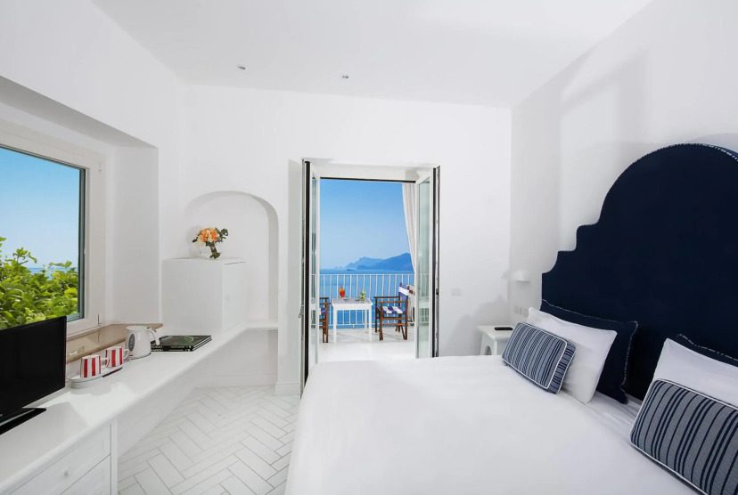 Bright white bedroom with blue accents and a large bed. Open double doors lead to a balcony with a stunning ocean view, evoking a serene, relaxing atmosphere.