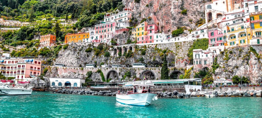 Colorful cliffside houses and lush greenery overlook a vibrant blue sea in an Italian coastal town. Boats gently float, creating a tranquil scene.