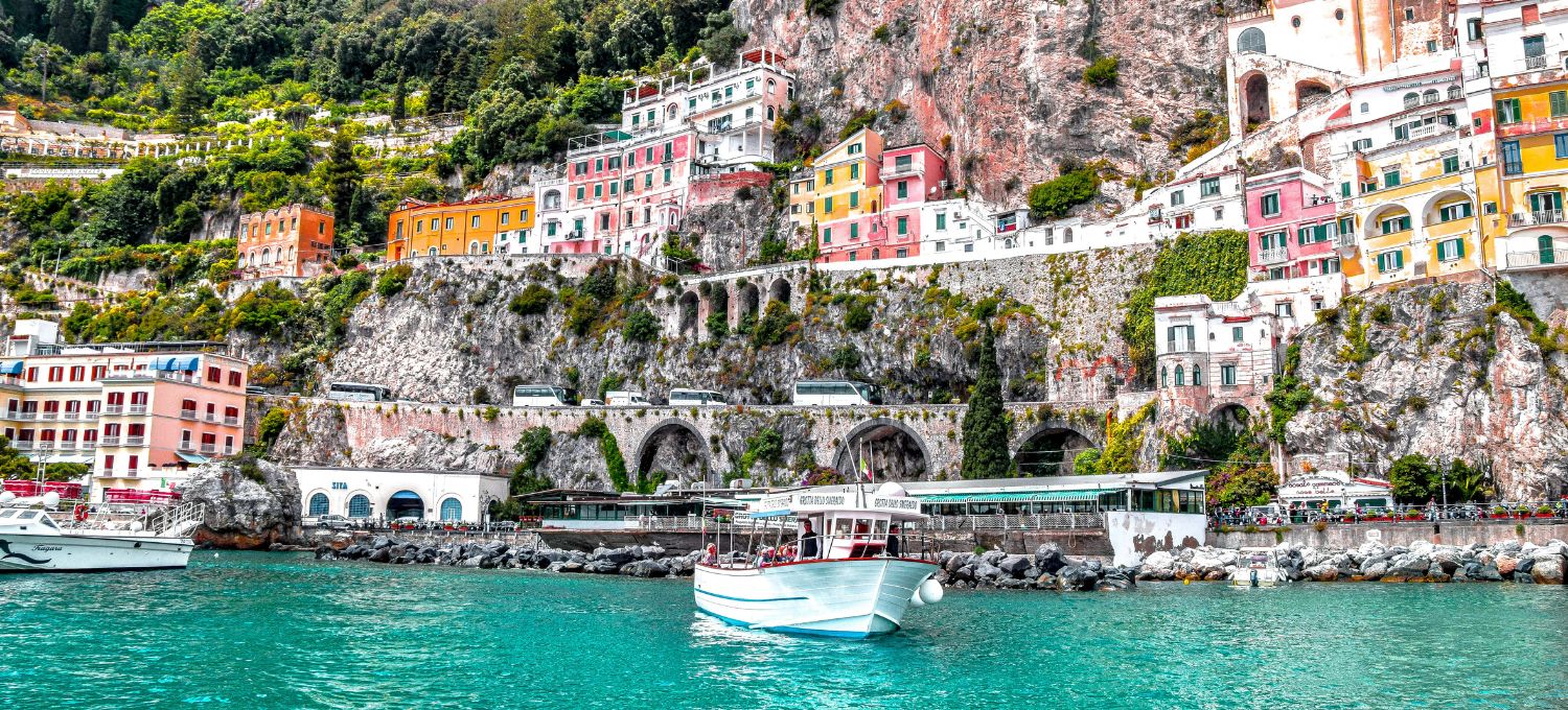 Colorful cliffside houses and lush greenery overlook a vibrant blue sea in an Italian coastal town. Boats gently float, creating a tranquil scene.