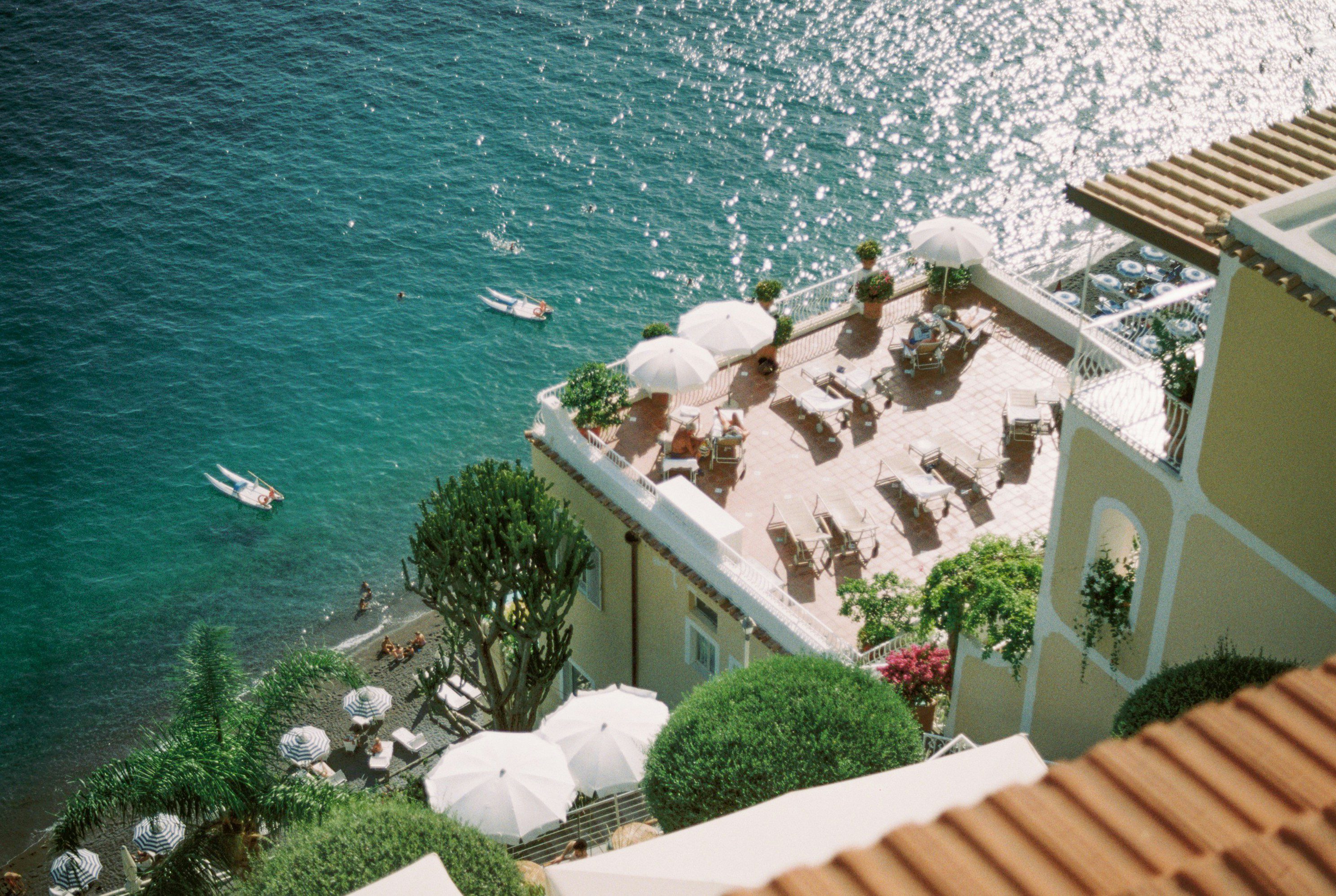 Aerial view of a seaside terrace with white parasols, chairs, and potted plants. The clear turquoise sea sparkles in the sunlight, creating a serene and inviting atmosphere.