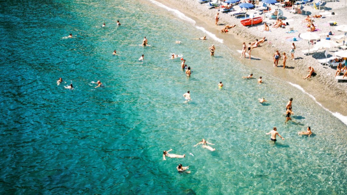 Aerial view of a crowded beach with people swimming in clear turquoise water. The sandy shore is lined with sunbathers and colorful umbrellas.