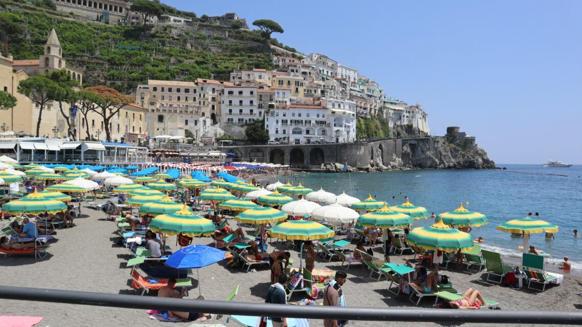 A vibrant beach scene with colorful umbrellas, sunbathers, and turquoise water. In the background, charming hillside buildings overlook the sea.