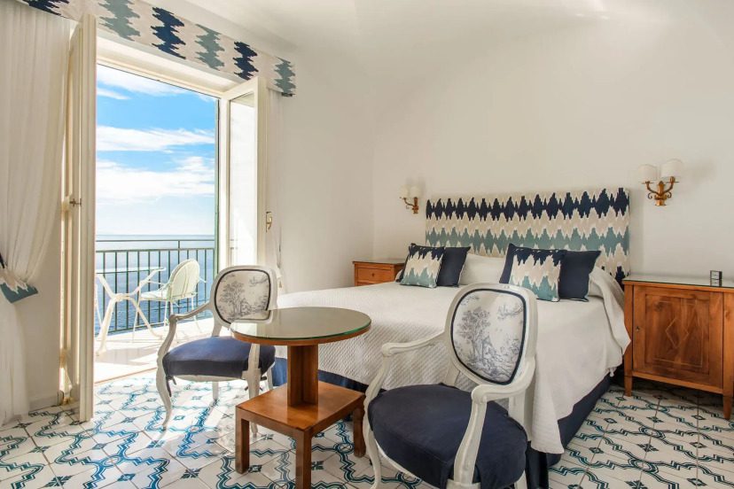 A bright, elegant bedroom with blue and white geometric patterns. A bed with matching pillows faces a glass table and chairs. An open balcony shows a sea view.