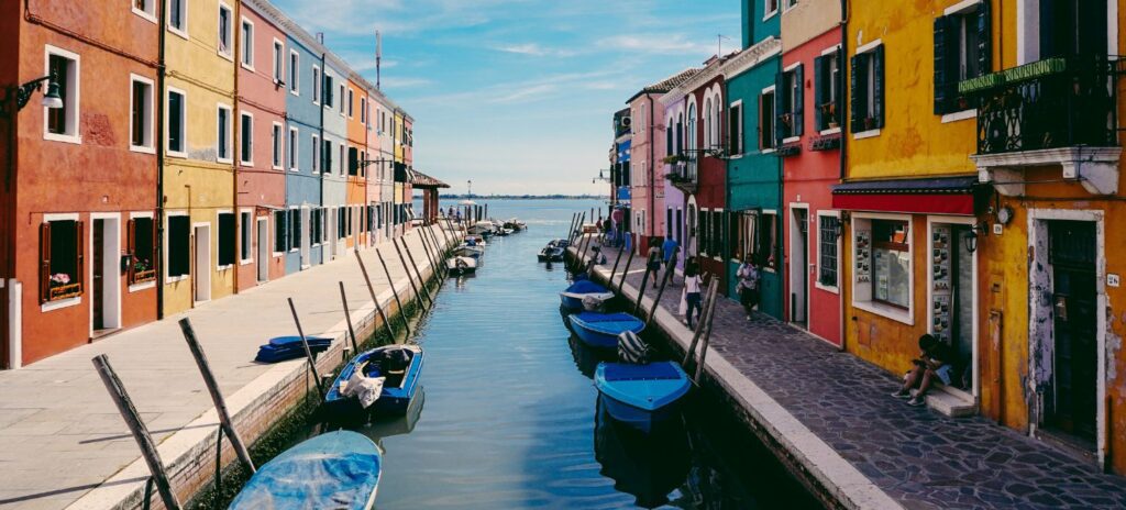 A vibrant canal scene in Burano, Italy, flanked by colorful buildings. Blue boats float on calm water under a clear sky, evoking a tranquil, lively atmosphere.