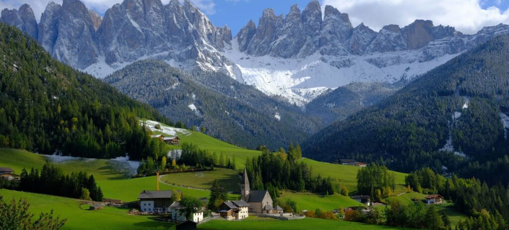 A serene valley village with quaint houses and a church is nestled in lush green hills, backed by towering snow-capped mountains under a partly cloudy sky.