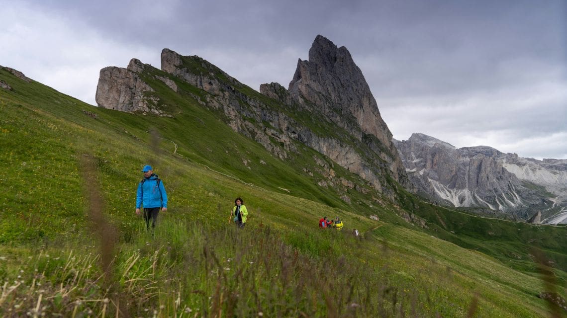 Hikers in vibrant jackets walk along a lush, green mountainside, under a dramatic, overcast sky. A jagged mountain peak looms in the background.
