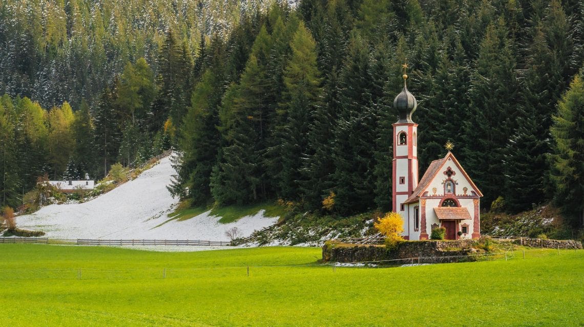 A quaint church with a steep, conical spire sits amid lush green fields and a forested mountain dusted with snow, evoking tranquility and isolation.