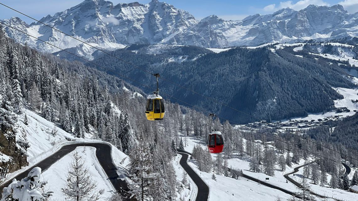 A snowy mountain landscape featuring yellow and red cable cars above winding roads, surrounded by snow-laden trees and majestic peaks under a clear sky.