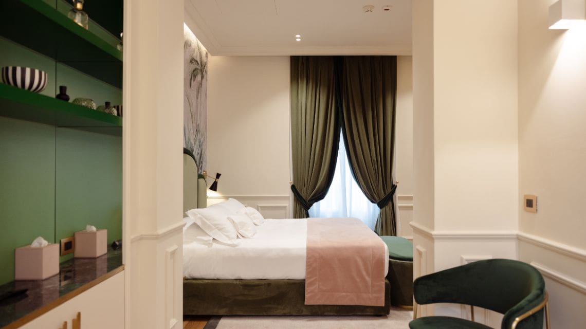 Elegant hotel room with neutral tones, featuring a plush bed with white and pink bedding. Olive curtains frame a window, creating a cozy ambiance.