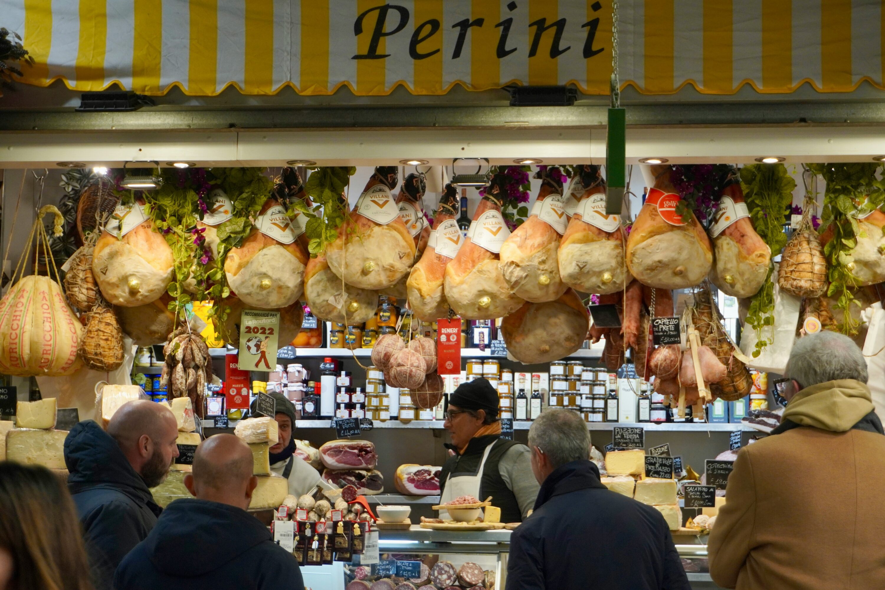 A bustling Italian deli showcases hanging cured meats and cheeses under warm lighting. Customers browse while a vendor assists, creating a lively and inviting atmosphere.