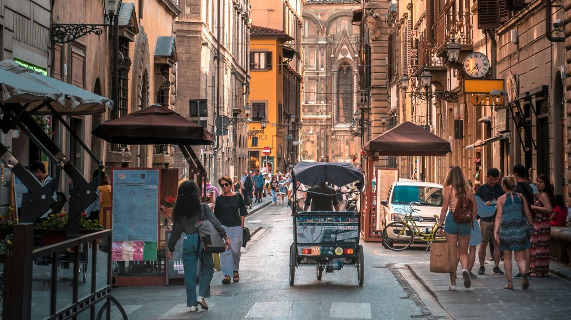 A bustling European street with people walking and a bike taxi in the center. Historic buildings line the narrow road, creating a lively, vibrant atmosphere.