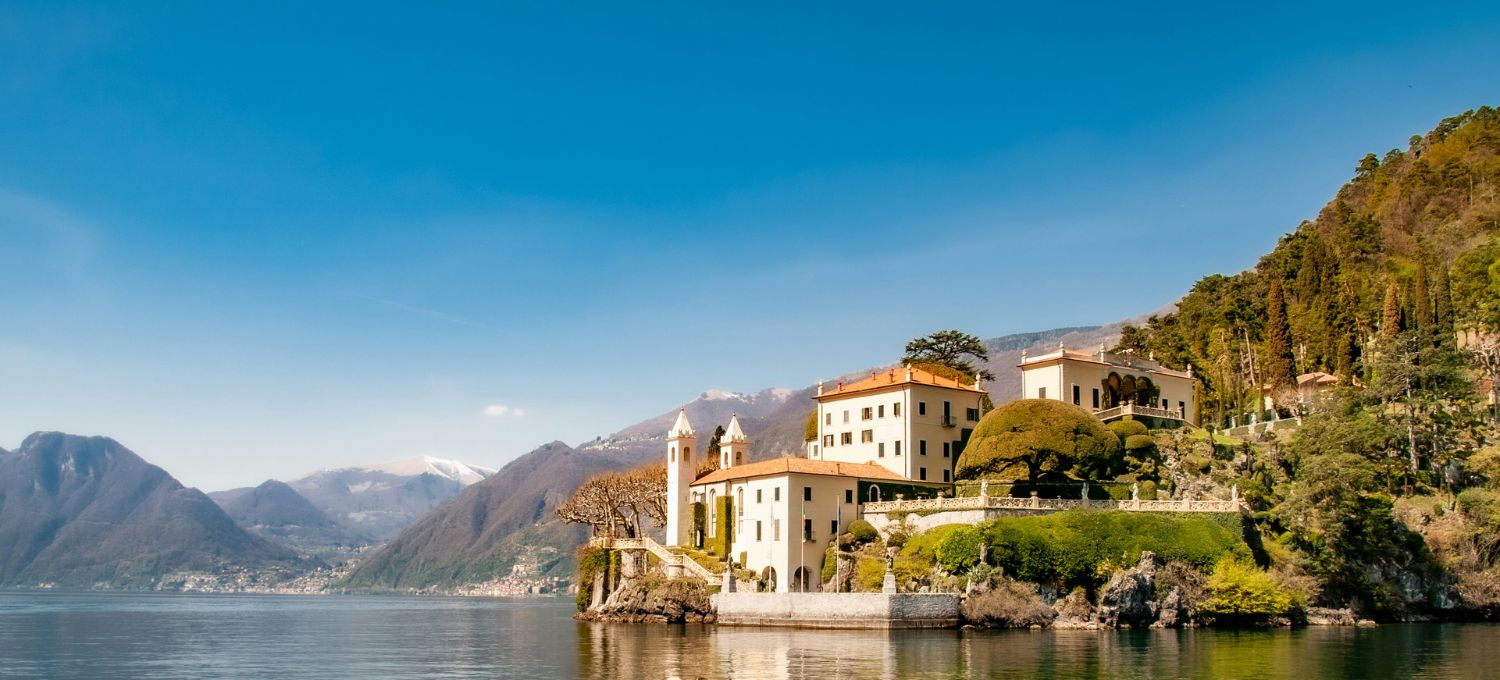 A picturesque villa perches on a lush, rocky outcrop by a serene lake. Surrounded by verdant trees and distant mountains under a clear, blue sky.