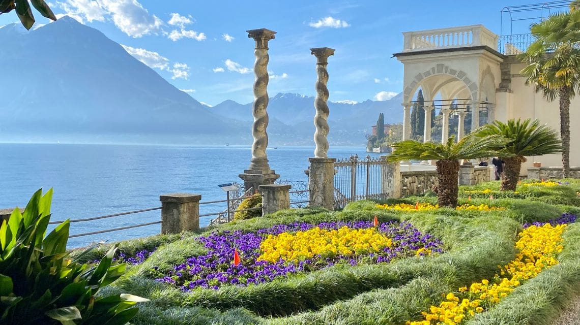 Elegant garden beside a serene lake, with vibrant yellow and purple flowers, twisted stone columns, and mountain backdrop under a clear blue sky.