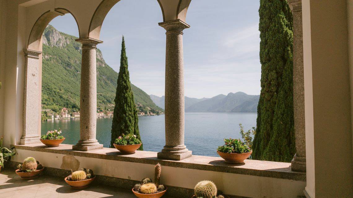 Stone columns frame a scenic view of a serene lake and distant mountains. Terracotta pots with lush plants and cacti line the sunlit terrace.