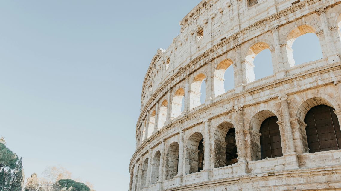 The image shows the Colosseum's exterior in Rome, Italy, under a clear blue sky. Sunlight highlights the ancient arches, evoking a sense of historic grandeur.