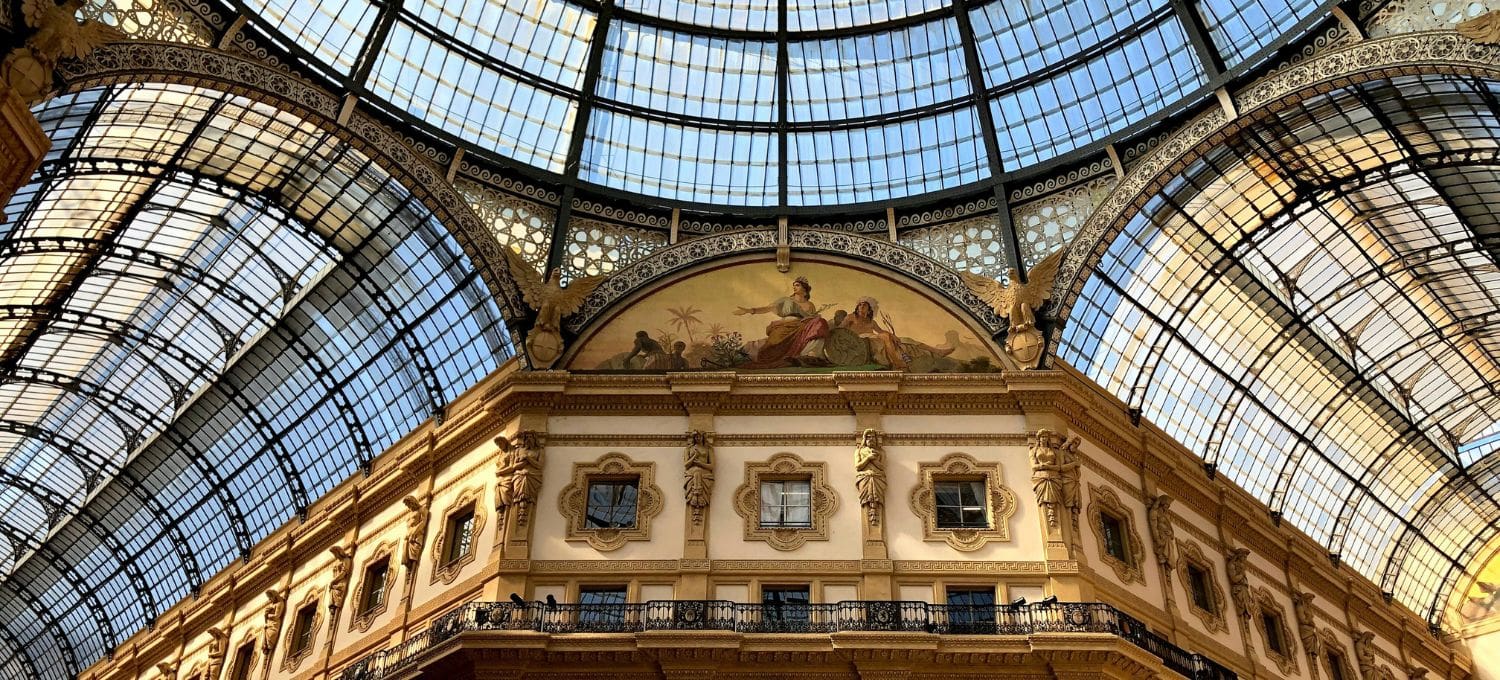 Ornate glass ceiling of a historic arcade with arching ironwork. Below, classical frescoes and detailed stonework evoke elegance and grandeur.