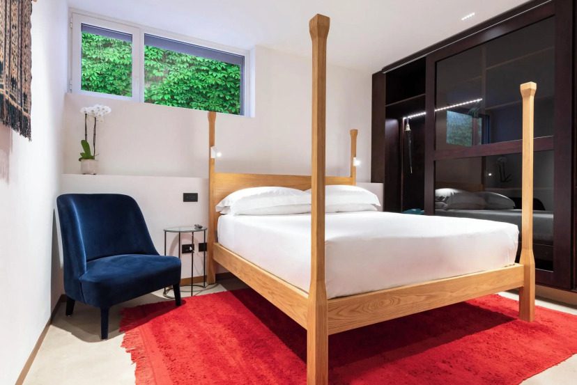Modern bedroom with a wooden four-poster bed on a vibrant red rug. A blue armchair and orchid adorn the room, while greenery is visible through a window. Calm ambiance.