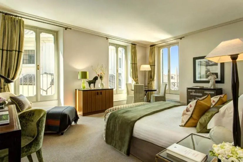 Bright hotel room with a double bed and green accents, featuring large windows, elegant decor, and cozy seating. A relaxed, inviting atmosphere.