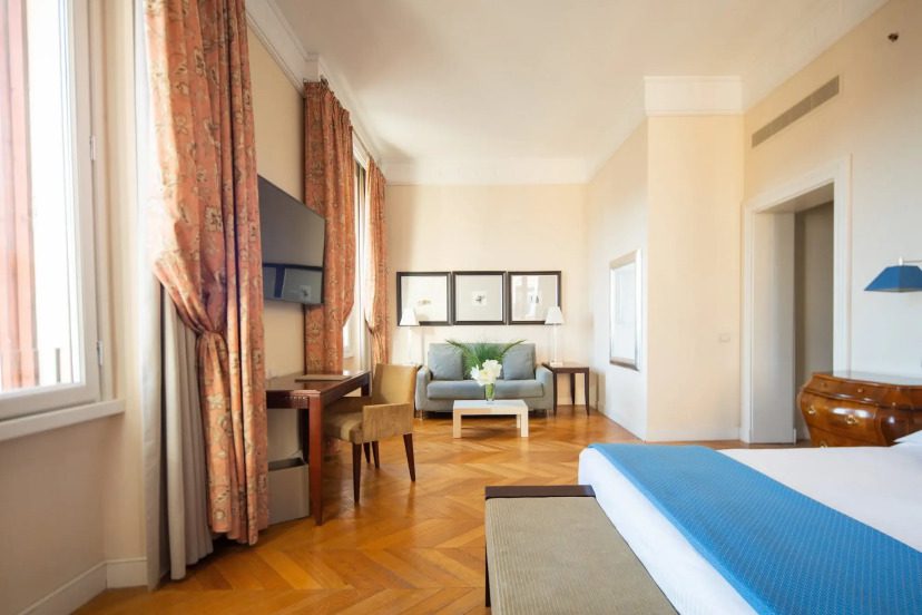 Elegant hotel room with wood flooring and soft peach walls. Features a blue-accented bed, a cozy seating area, desk, and large windows with floral curtains.