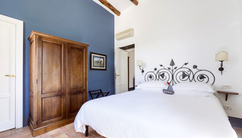 Minimalist bedroom with a white bed, ornate metal headboard, and wooden wardrobe. Blue accent wall adds contrast. Warm and inviting atmosphere.