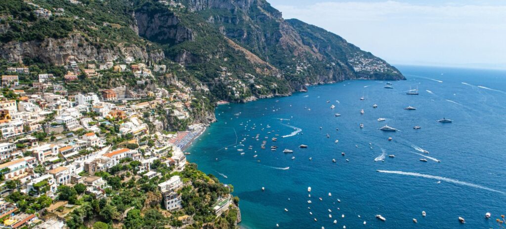 Aerial view of a coastal town with colorful buildings on a steep cliffside, overlooking a sparkling blue sea filled with boats. The scene is vibrant and serene.