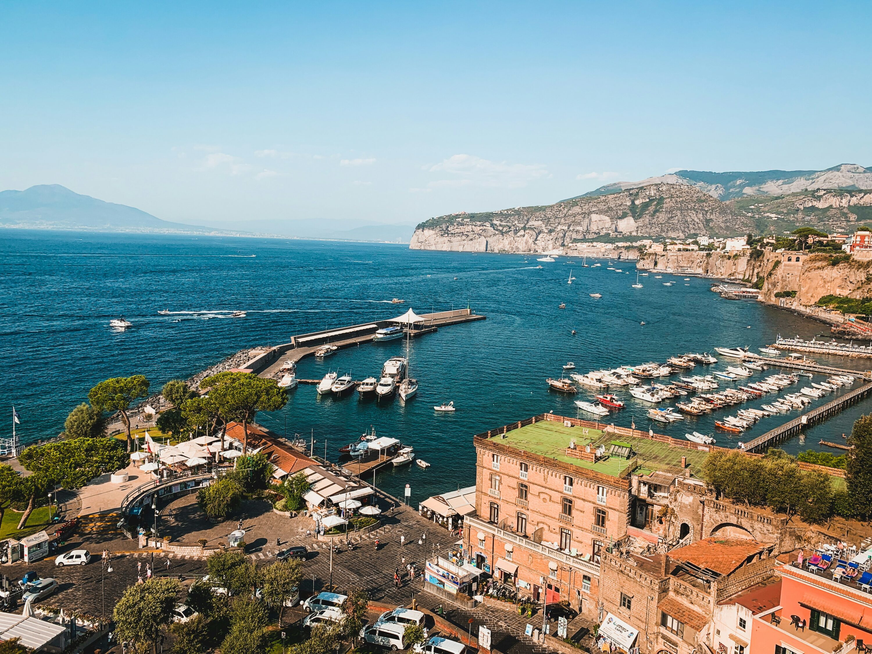 A scenic view of a coastal town with a marina full of boats, clear blue sea, and distant mountains. The atmosphere is bright and serene under a sunny sky.