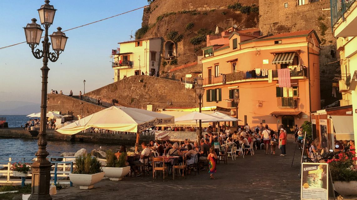 Outdoor cafe in a coastal village at sunset. Patrons sit under white umbrellas near colorful buildings with hanging laundry. Warm, relaxed atmosphere.