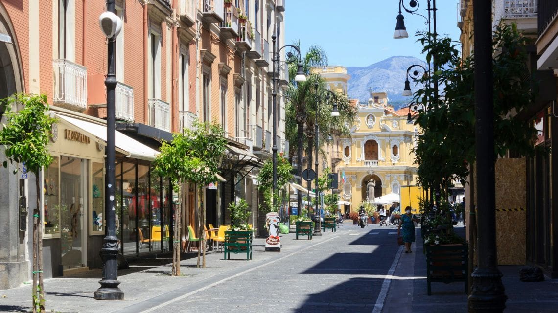 A sunlit, narrow street with charming buildings on both sides, lined with small trees and outdoor seating. People leisurely stroll towards a distant, ornate facade.