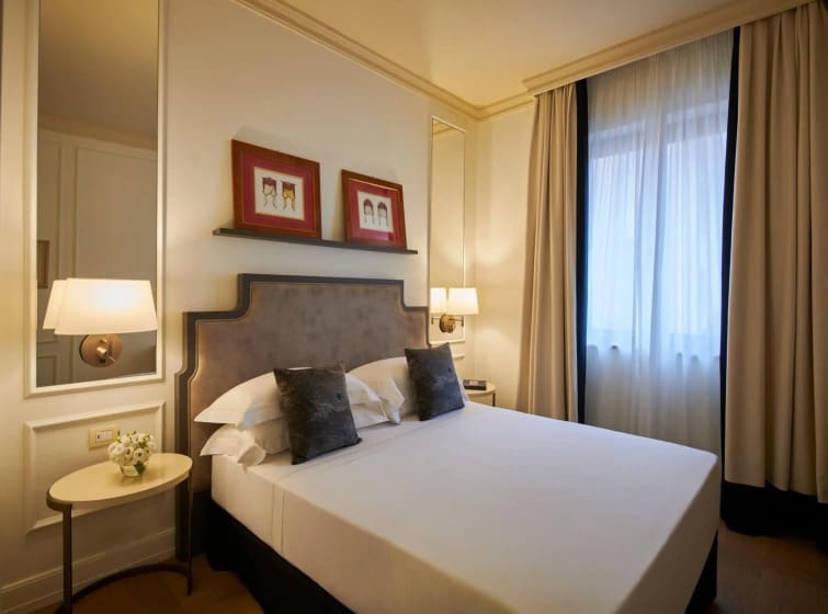 A cozy hotel room with a neatly made double bed, two plush pillows, elegant wall sconces, art in red frames above, and curtains softly lit by daylight.