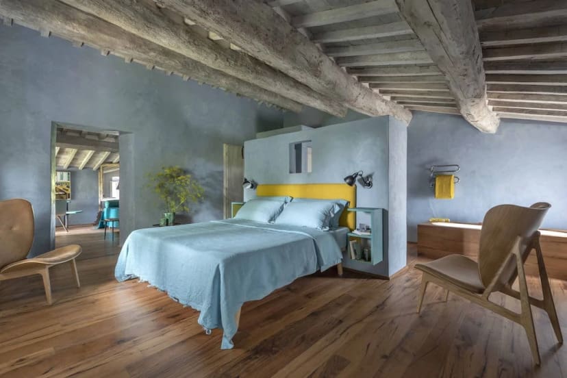 Spacious bedroom with rustic wooden beams and floor, featuring a bed with light blue bedding, yellow accents, modern chairs, and soft gray walls. Cozy and serene.