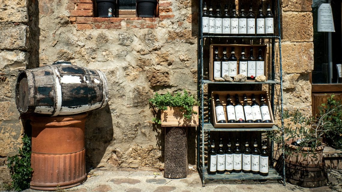 A rustic outdoor setting features a weathered barrel on a pedestal, a wooden rack displaying wine bottles against a stone wall, and potted plants. Warm, nostalgic ambiance.