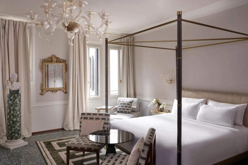 Elegant hotel room with a canopy bed, white bedding, and plush chairs. A chandelier hangs from the ceiling. Light filters through tall windows, creating a serene ambiance.