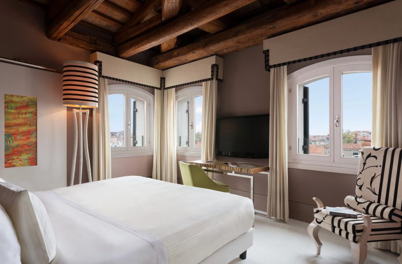 Elegant bedroom with a beamed ceiling, three arched windows, a striped lamp, and a TV on a desk. Features cozy bedding, patterned chair, calm ambiance.