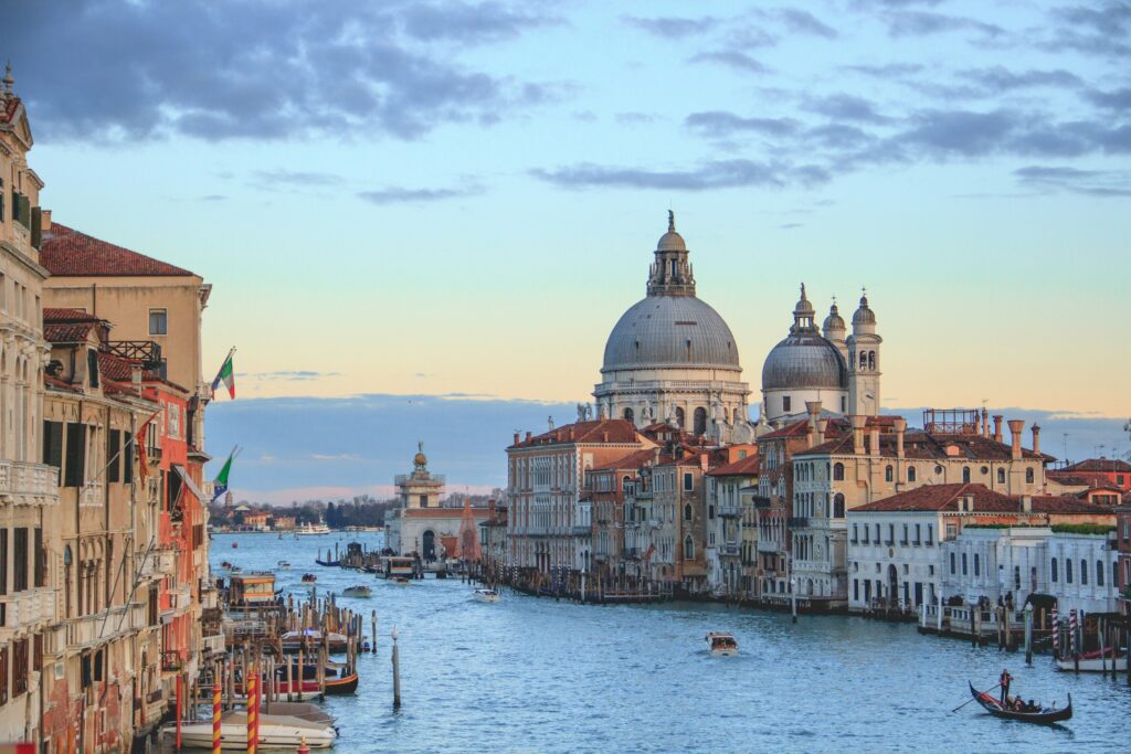 Scenic view of Venice's Grand Canal at sunset, featuring historic buildings with domes and flags. Gondolas and boats glide on calm water, creating a serene atmosphere.