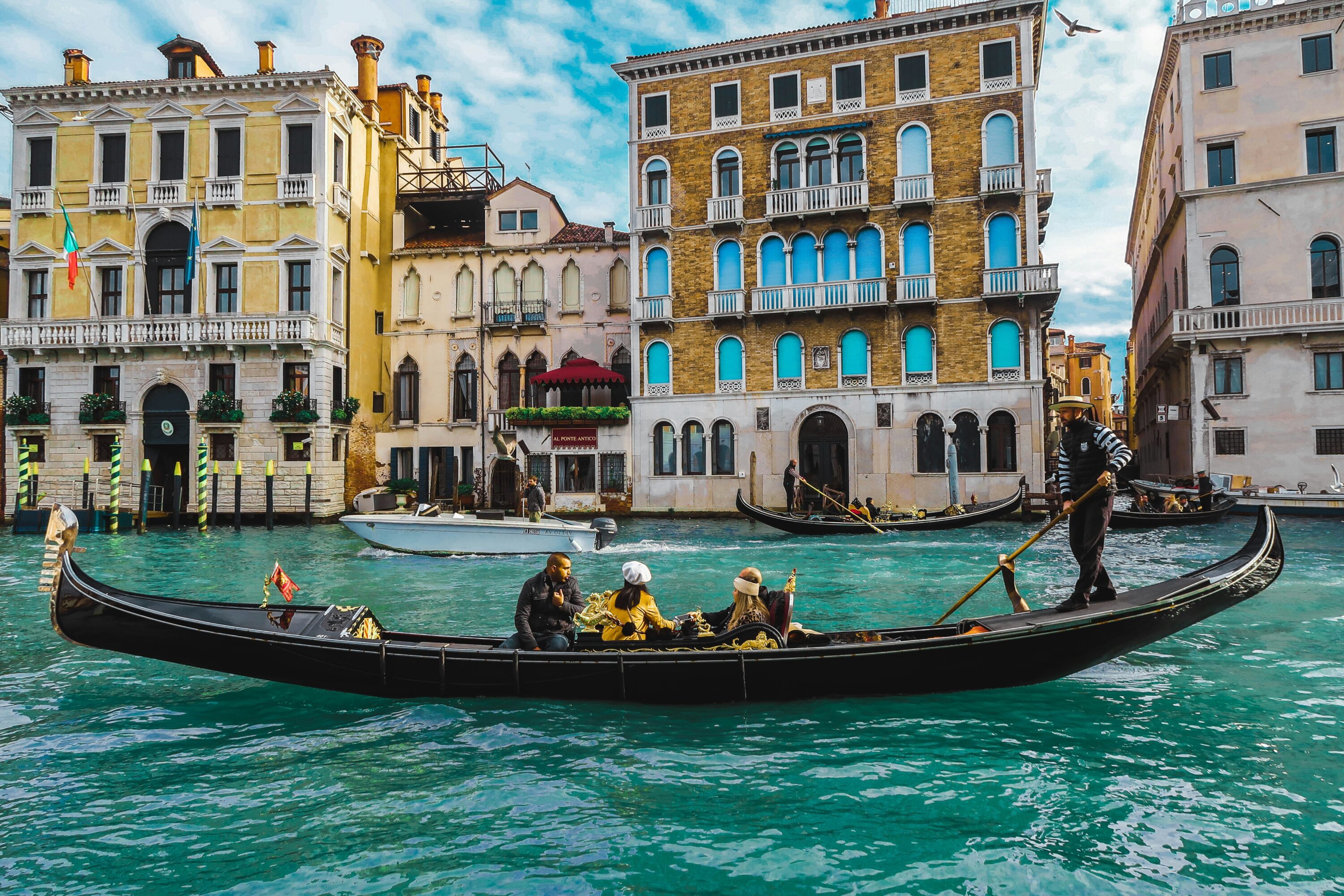 Gondola on Venetian canal with three passengers, steered by gondolier. Vibrant historic buildings and clear blue sky in background. Relaxed, scenic vibe.