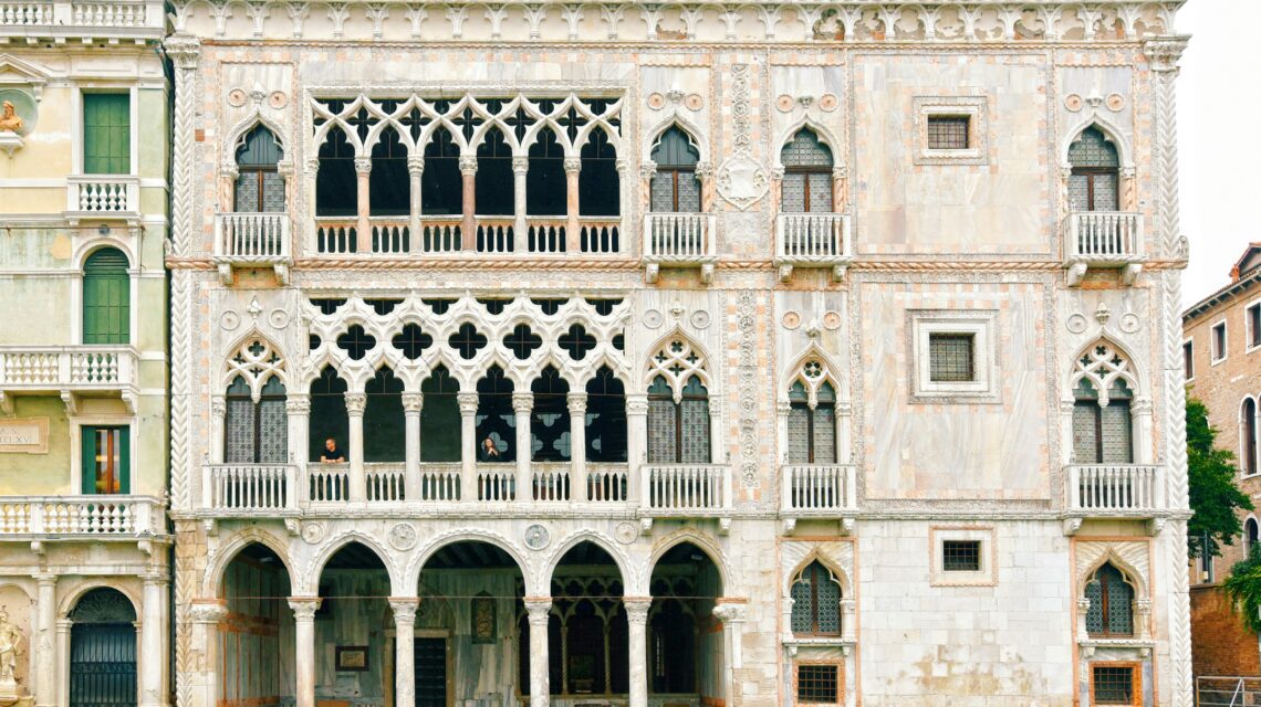 An ornate Venetian palace with intricate Gothic architecture, featuring arched windows, decorative stonework, and reflections in the canal below.