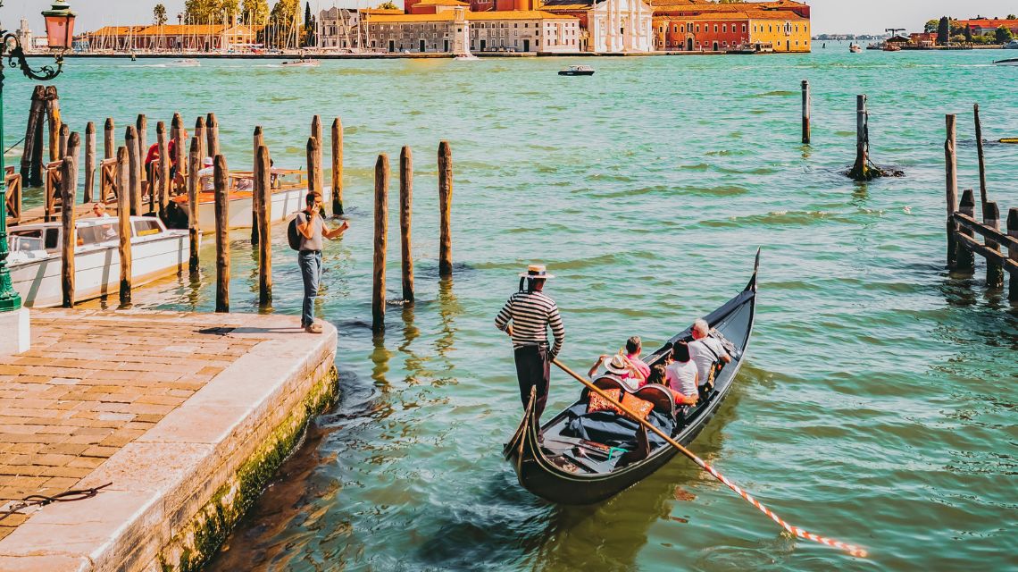 A gondola with a gondolier and passengers glides through turquoise Venetian waters, with San Giorgio Maggiore island's historic church in the sunny background.