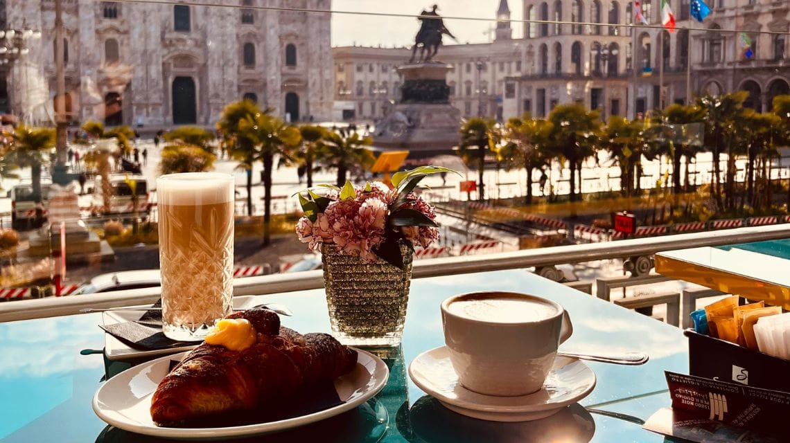 A cup of coffee, croissant, and latte on a table with a flower vase, overlooking a city plaza with a statue, palm trees, and historic architecture.