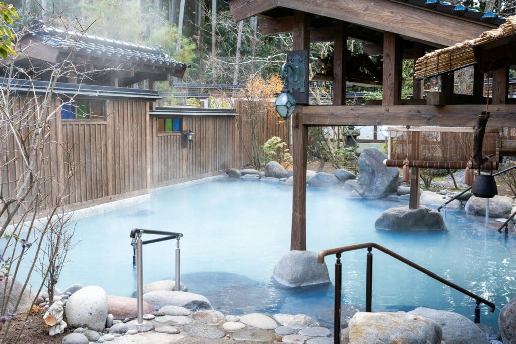 A serene outdoor hot spring bath surrounded by steam, wooden structures, and smooth stones. The scene conveys tranquility and relaxation.