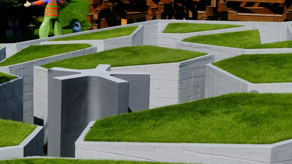 A winding gray stone maze with green grass tops, in the sunlight. A colorful, whimsical statue stands at the background, suggesting a playful atmosphere.