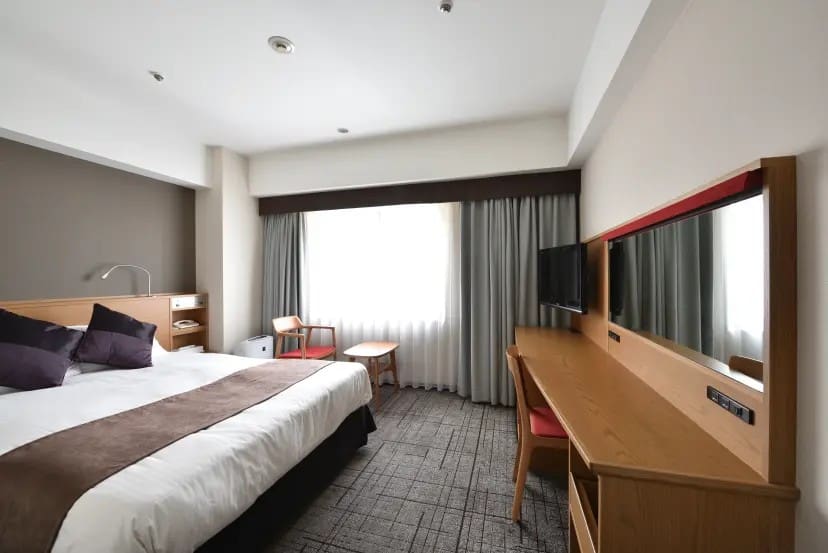 Modern hotel room with a large bed, white and brown bedding, purple pillows, beside a wooden desk, TV, and window with gray curtains, conveying a cozy feel.