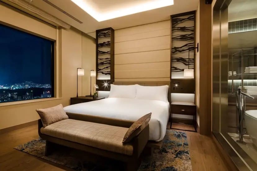 Elegant hotel room at night with a large bed, modern decor, and a view of a city skyline through a window. Warm lighting creates a cozy ambiance.