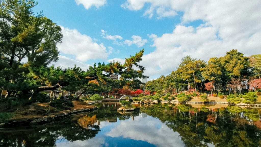 Tranquil Japanese garden scene with a serene pond reflecting lush trees and a small stone bridge under a blue sky with scattered clouds.