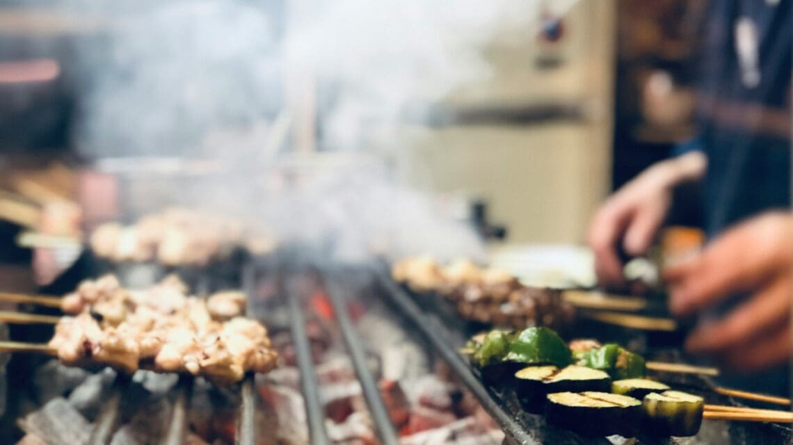 Skewers of grilled vegetables and meat over open flames fill the foreground with smoke. A person tends to the grill, creating a lively, bustling scene.