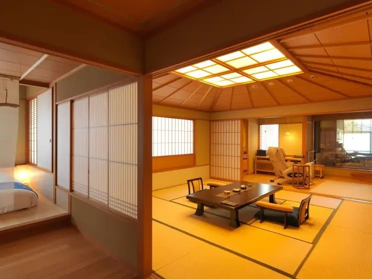 Traditional Japanese room with tatami mats, low wooden table, and shoji screens. Soft lighting creates a serene, inviting atmosphere. A bed is partially visible.