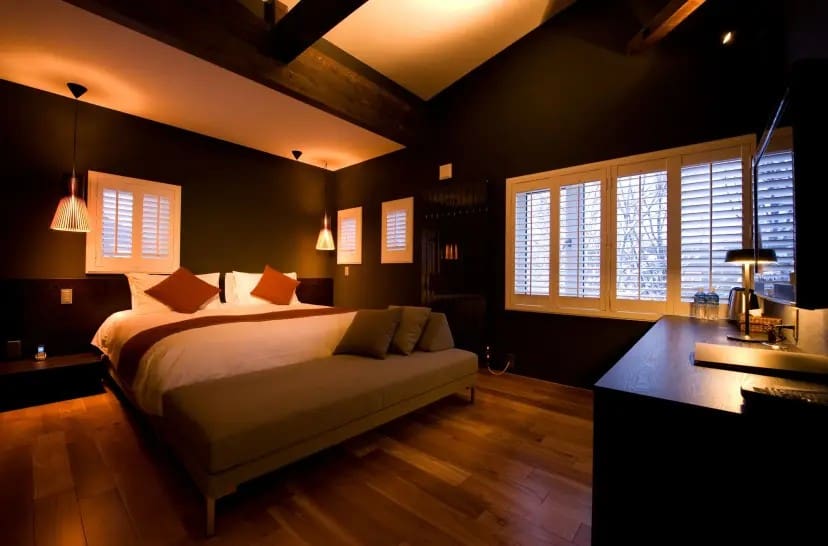 Cozy bedroom with warm lighting, featuring a large bed with brown and white bedding, pendant lights, wooden floors, and shuttered windows.